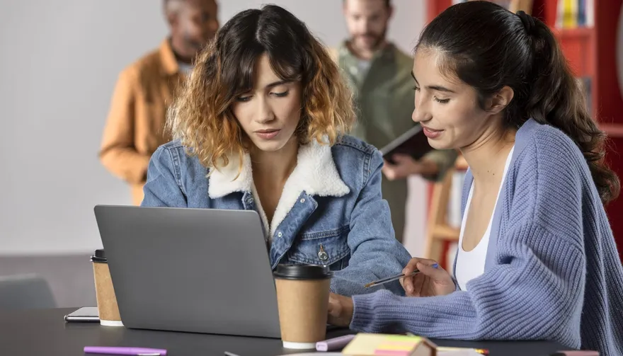 Two colleagues sitting together at a laptop in a busy workspace, discussing what is on screen.