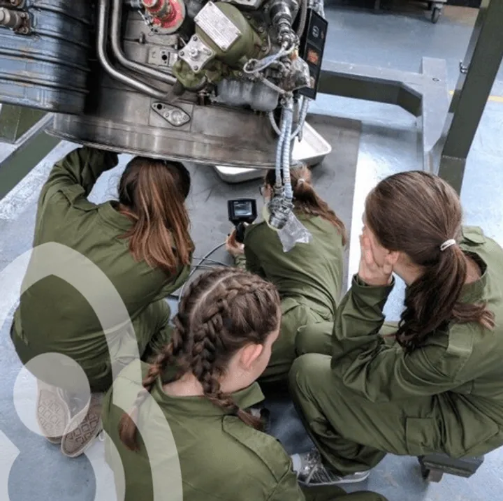 Apprentices in overalls gathered around a jet engine, inspecting it with a borescope camera.